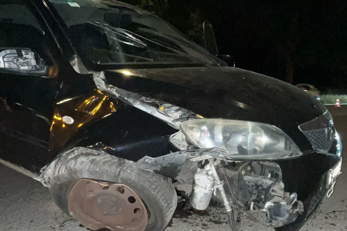 Das Bild zeigt ein schwer beschädigtes schwarzes Auto mit kaputtem Vorderreifen und eingedrückter Front auf einer Straße bei Nacht. Unfällelemente sind erkennbar, das Auto steht still.