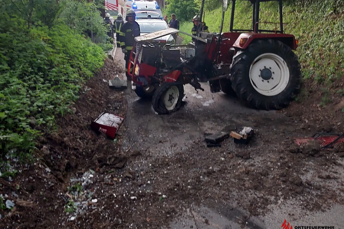Ein roter Traktor steht stark beschädigt auf einem schmalen Waldweg. Mehrere Einsatzkräfte sind anwesend, sowie ein Rettungsfahrzeug im Hintergrund.