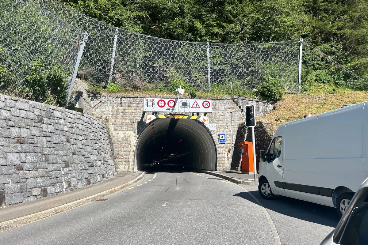 Ein weißer Lieferwagen steht vor einem Tunnel, daneben ist eine Ampel und verschiedene Verkehrsschilder sichtbar. Der Tunnel ist von einer steinernen Mauer umgeben.