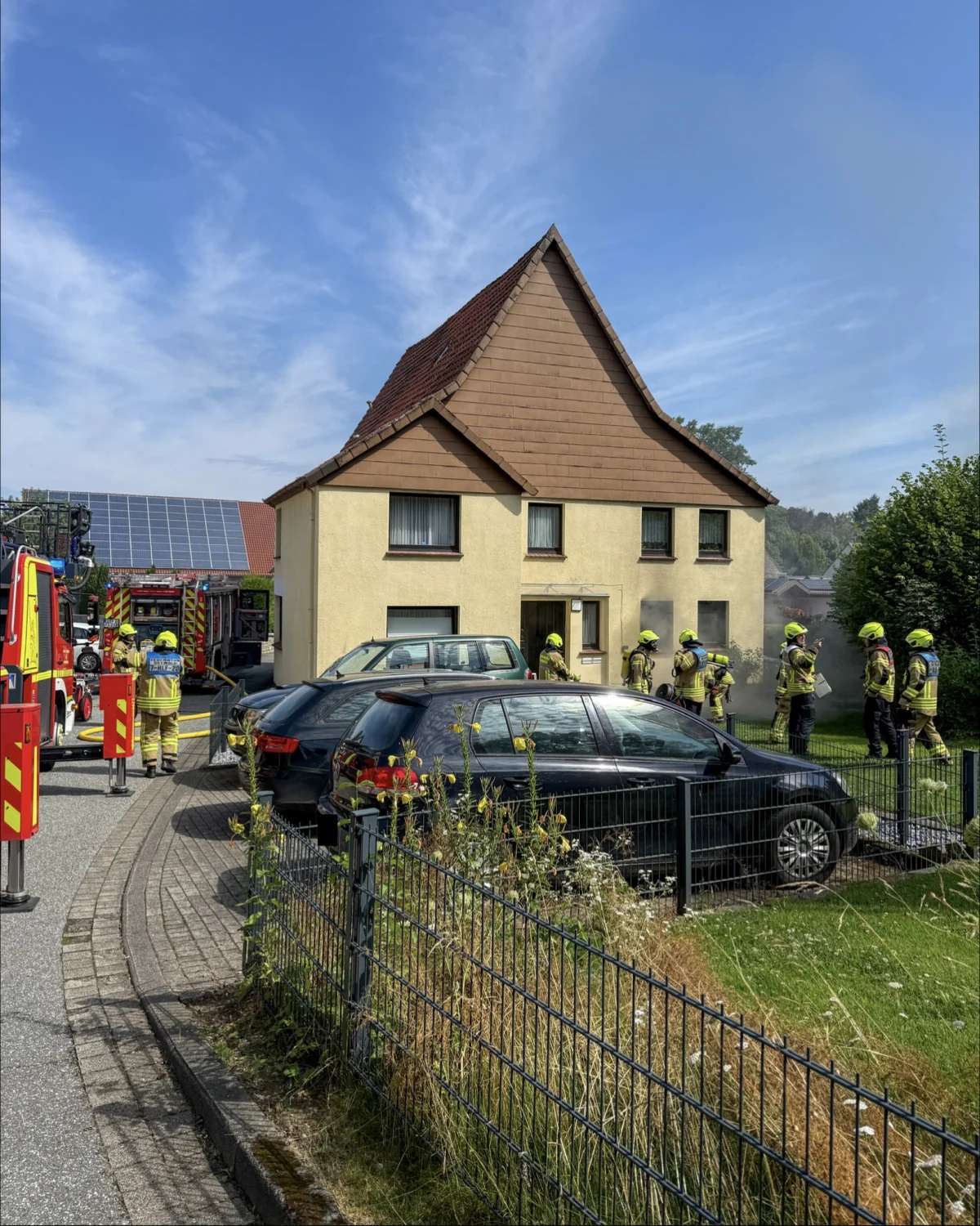 Ein Wohnhaus mit Feuerwehrleuten, die offenbar auf Rauchentwicklung reagieren. Mehrere Fahrzeuge sind im Vordergrund geparkt.