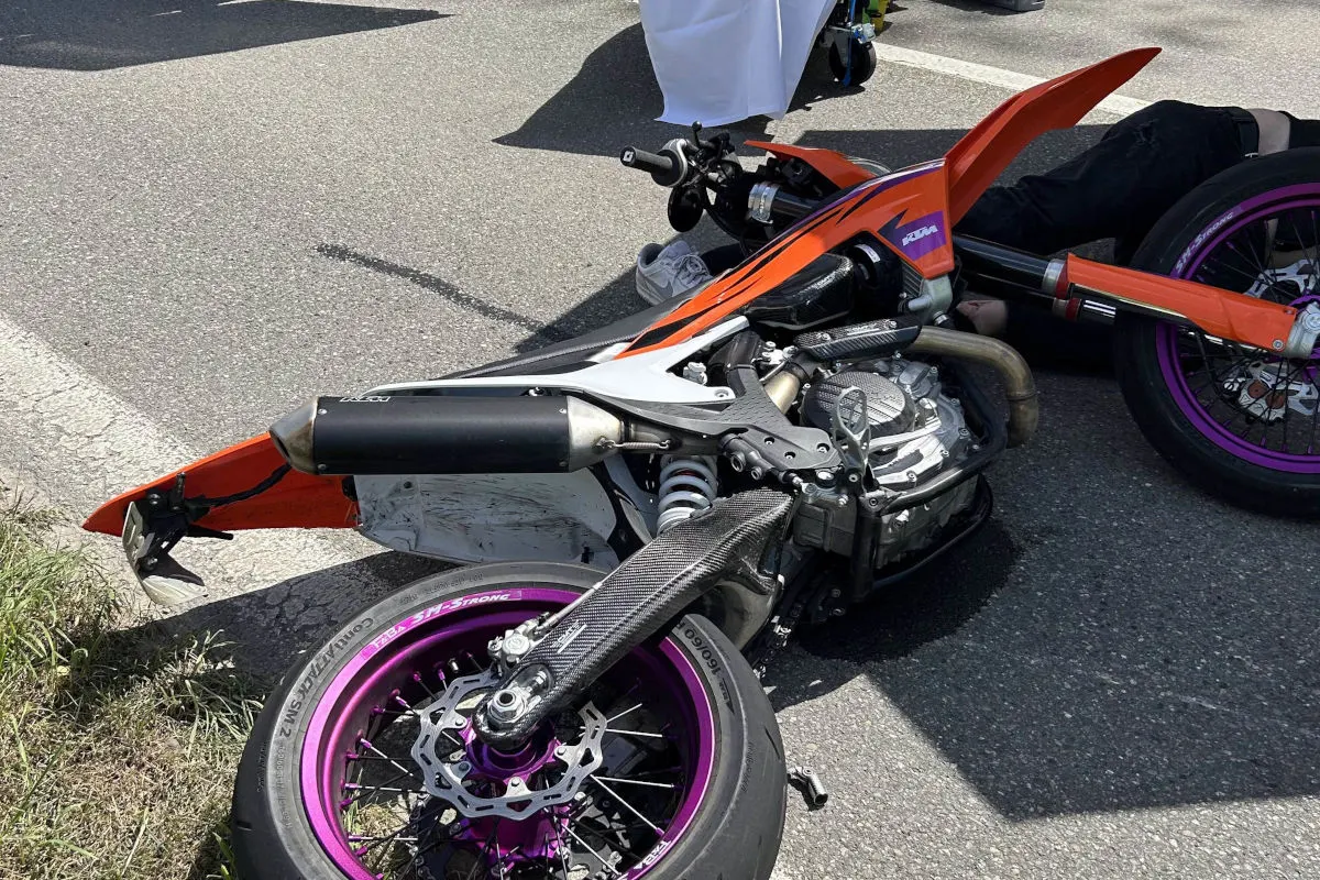Ein orangefarbenes Motorrad liegt gestürzt auf der Straße mit sichtbaren Schäden. Ein Helm und ein Schuh sind in der Nähe zu sehen.