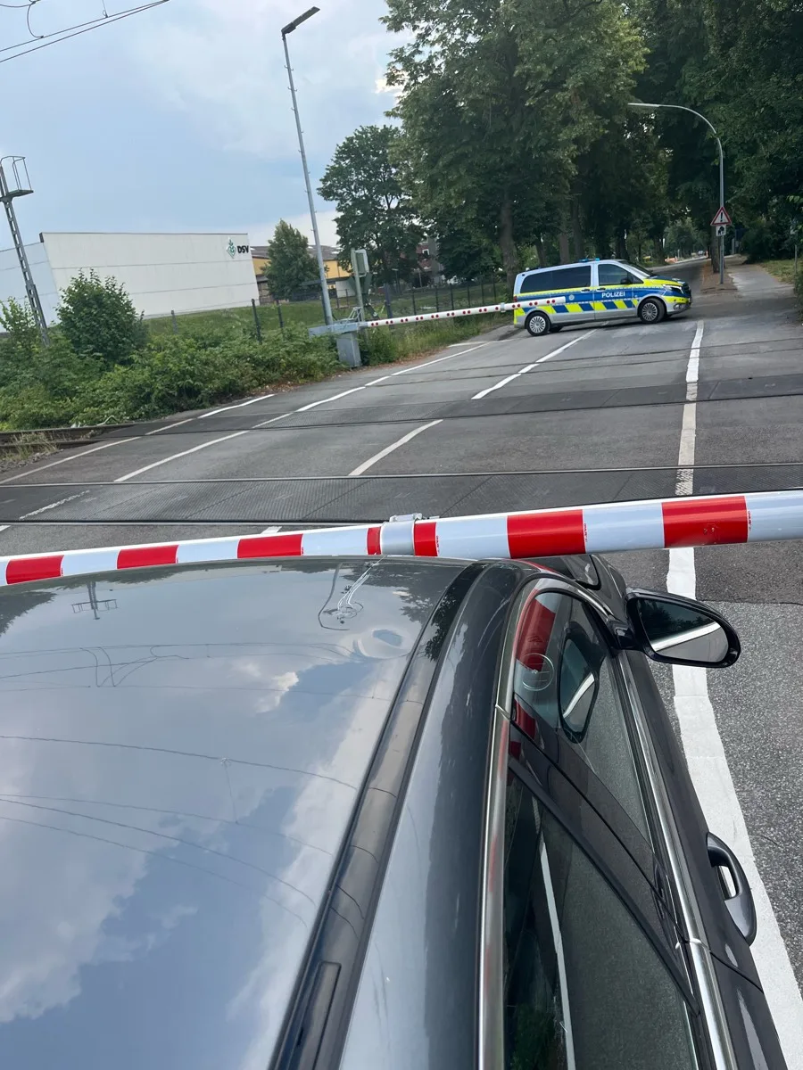 Ein Polizeifahrzeug mit blauen und gelben Markierungen blockiert eine Straße. Im Vordergrund ist eine rot-weiße Absperrung zu sehen.