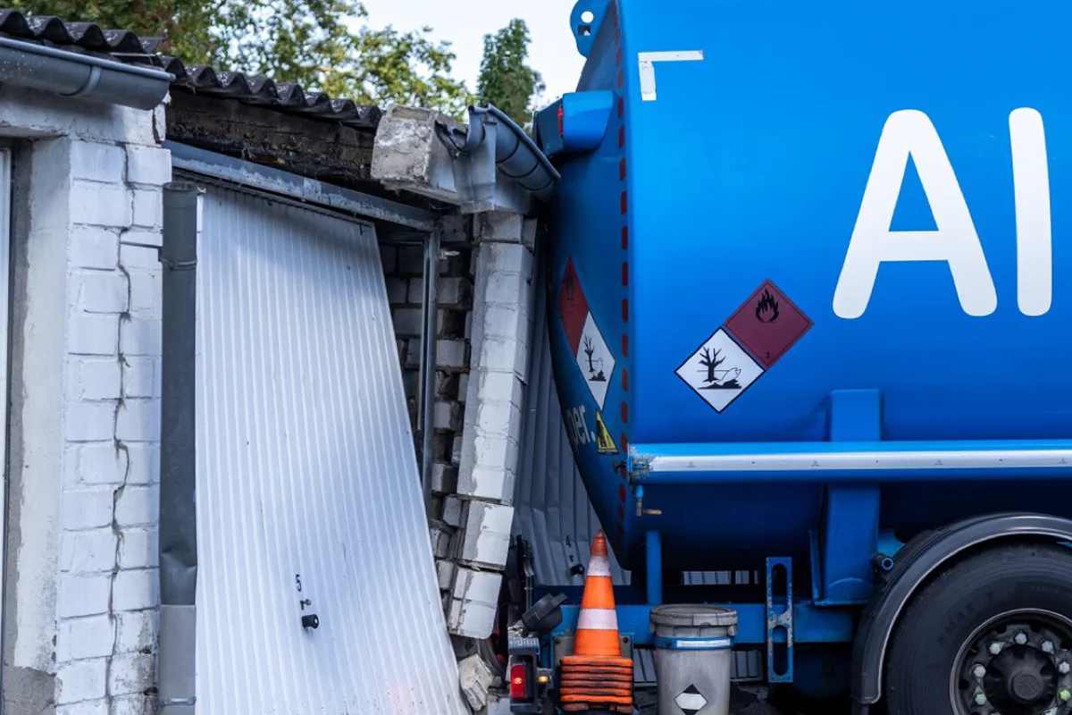 Ein blauer Gefahrgut-Lkw ist in eine Garage gefahren, wodurch die Wand und das Tor stark beschädigt wurden. Warnsymbole auf dem Lkw zeigen Gefahrenstoffe an.