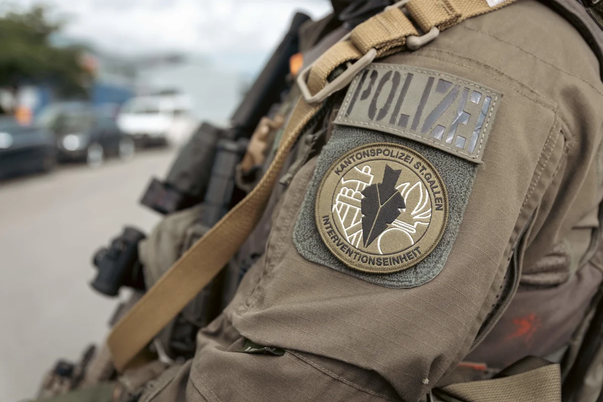 Das Bild zeigt das Abzeichen der Kantonspolizei St. Gallen auf der Uniform eines Polizisten. Im Hintergrund sind unscharf geparkte Autos zu sehen.
