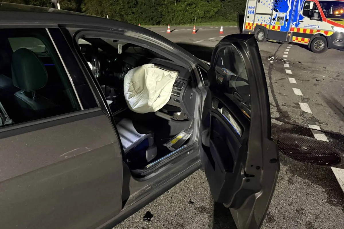 Ein geöffneter Airbag ist im Inneren eines dunklen Autos zu sehen. Im Hintergrund steht ein Krankenwagen und es liegen Trümmerteile auf der Straße.