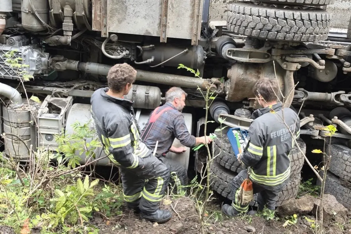 Ein Lastwagen liegt auf der Seite, während Feuerwehrleute die Situation begutachten. Das Gelände ist bewachsen mit niedrigem Grün.