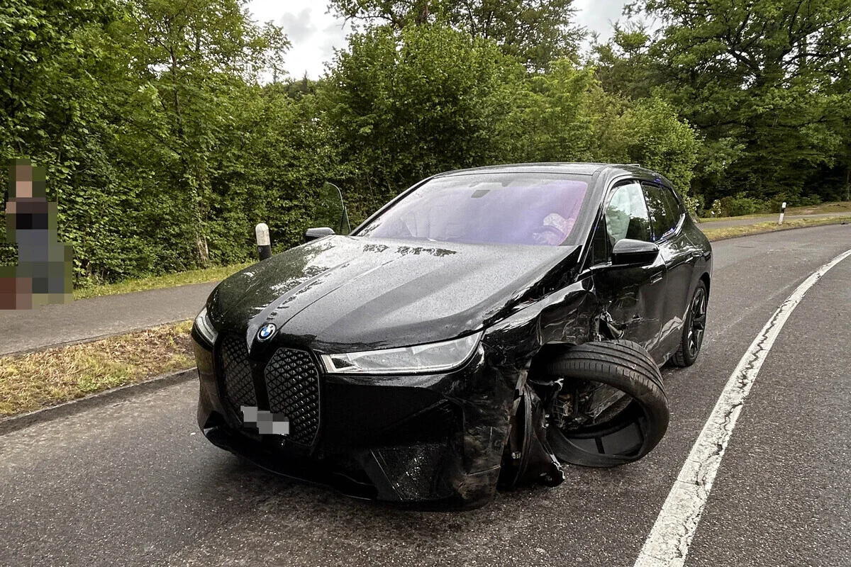 Ein schwarzer BMW mit stark beschädigtem Vorderrad steht auf einer nassen Landstraße. Im Hintergrund ist üppige Vegetation erkennbar.