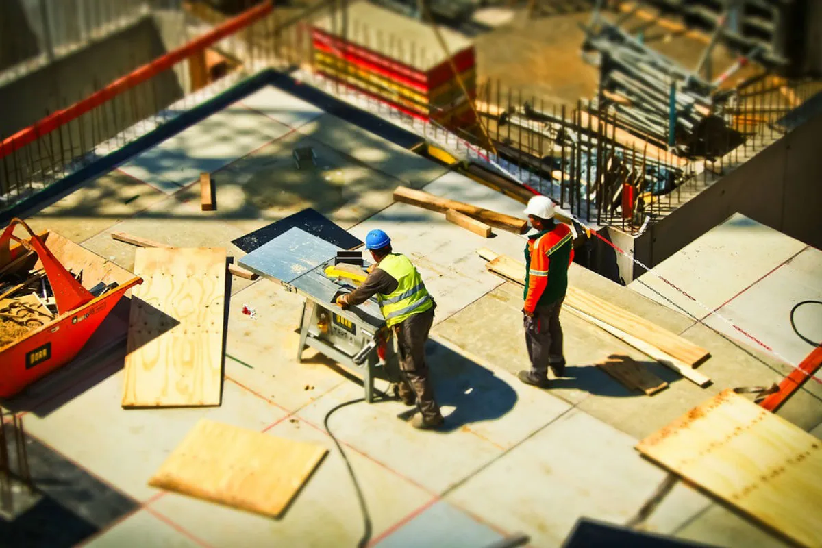 Die Aufnahme zeigt zwei Bauarbeiter auf einer Baustelle, einer mit gelber Warnweste arbeitet an einer Säge. Verschiedene Baumaterialien liegen herum.
