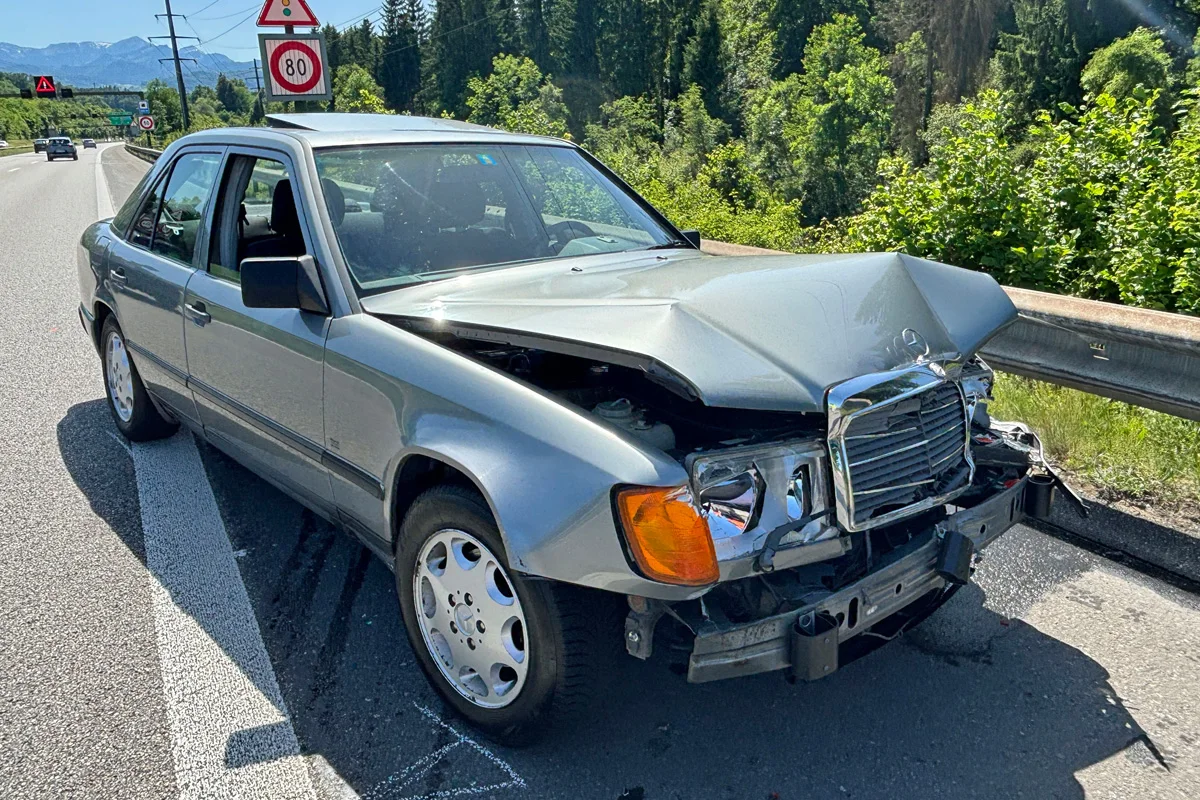 Ein silberner Mercedes mit stark beschädigter Front steht auf dem Seitenstreifen einer Autobahn. Im Hintergrund sind Bäume und Verkehrsschilder zu sehen.