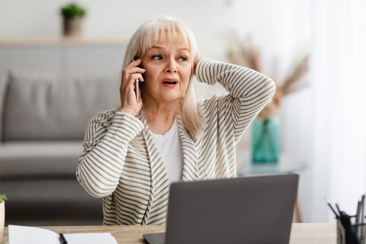 Eine ältere Frau mit weißen Haaren hält sich besorgt das Gesicht, während sie mit einem Smartphone telefoniert. Sie sitzt in einem hellen Raum vor einem Laptop.
