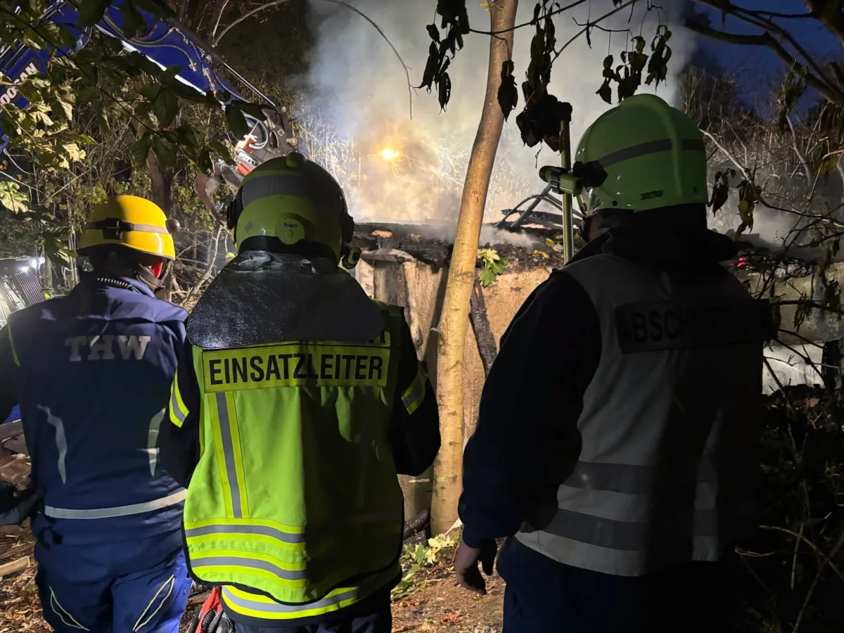 Drei Einsatzkräfte in Schutzkleidung stehen vor einem rauchenden Brandherd im Wald. Zwei tragen auffällige Jacken mit Aufschriften wie 'Einsatzleiter' und 'Abschnitt'.