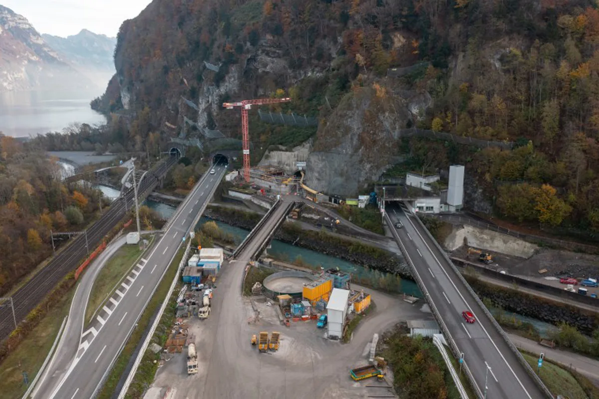 Luftaufnahme einer Straße mit zwei Tunnelöffnungen in einer bergigen Landschaft. Baustellenfahrzeuge und Kräne sind sichtbar.