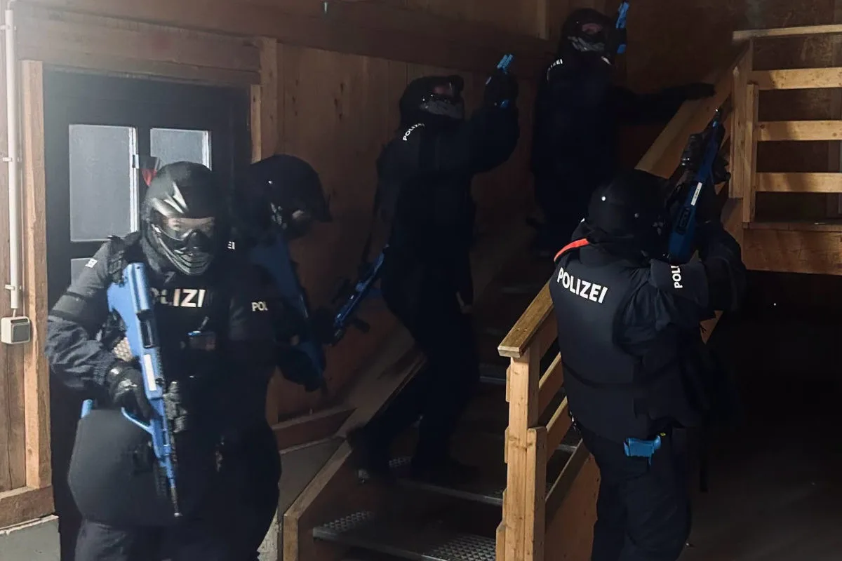 Eine Gruppe von Polizisten in vollständiger Schutzmontur und mit blauen Waffen marschiert eine hölzerne Treppe in einem Gebäude hinauf.