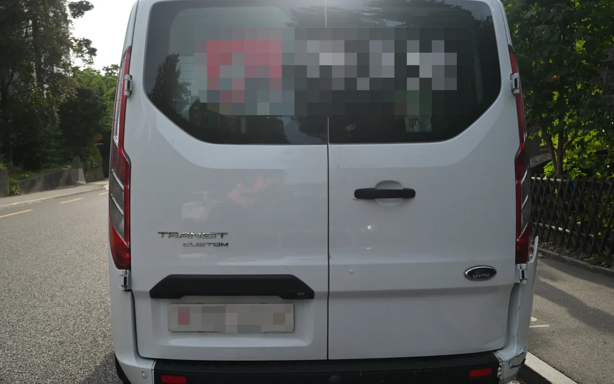 Die Rückseite eines weißen Ford Transit Custom zeigt sichtbare Beschädigungen an einer Straßenecke bei Tageslicht.
