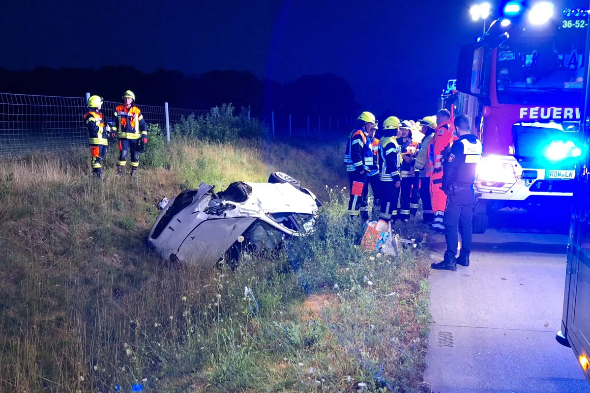 Ein weißes Auto liegt auf dem Dach in einem Straßengraben, während mehrere Feuerwehrleute und ein Polizist daneben stehen und den Unfallort absichern.