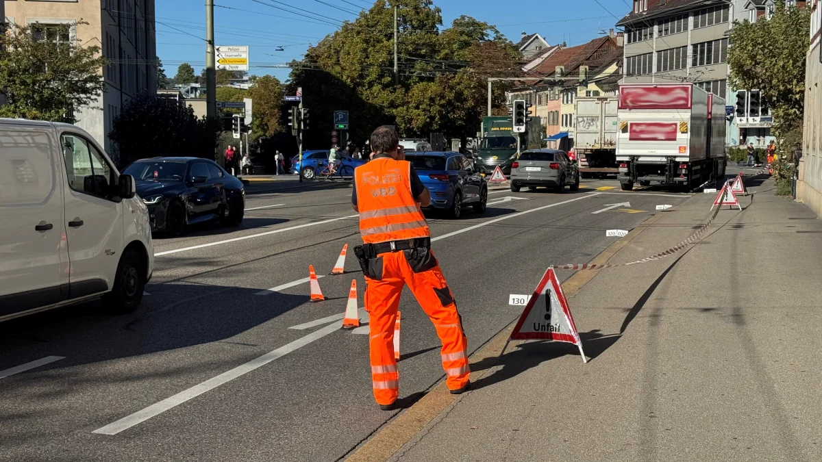 Ein Polizist trägt eine orangefarbene Warnweste und sichert eine belebte Straße mit Verkehrshütchen ab, während Autos vorbeifahren.