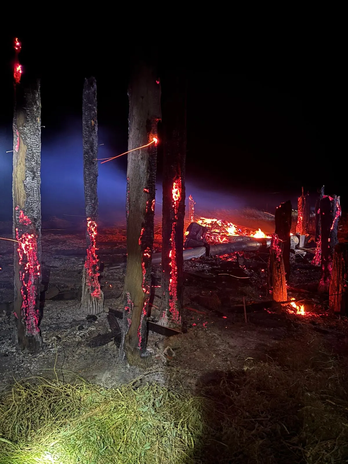 Mehrere hohe Holzpfosten stehen in Flammen auf einem dunklen Feld, umgeben von glühender Asche.