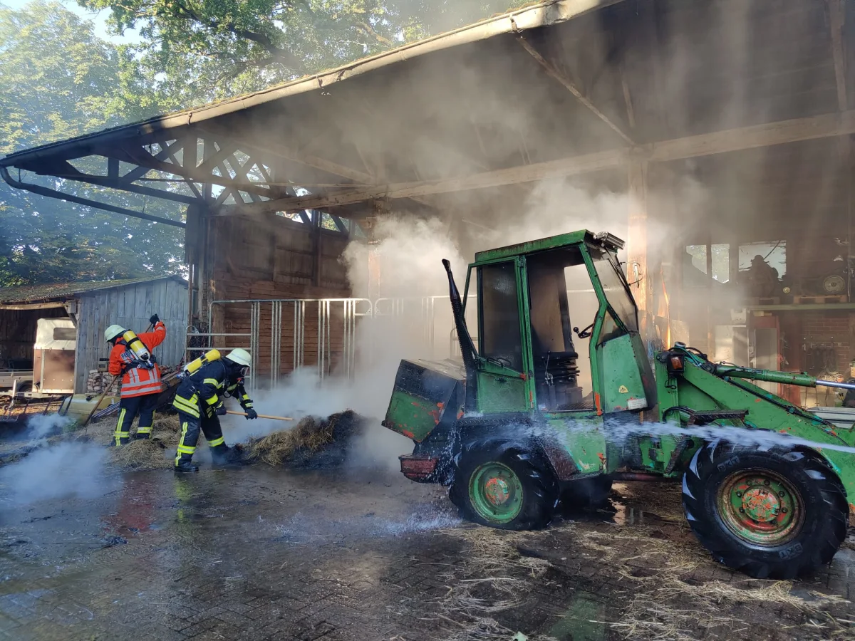 Zwei Feuerwehrleute bekämpfen ein Feuer in einer Scheune. Rauch steigt auf, und ein grünes landwirtschaftliches Fahrzeug steht daneben.