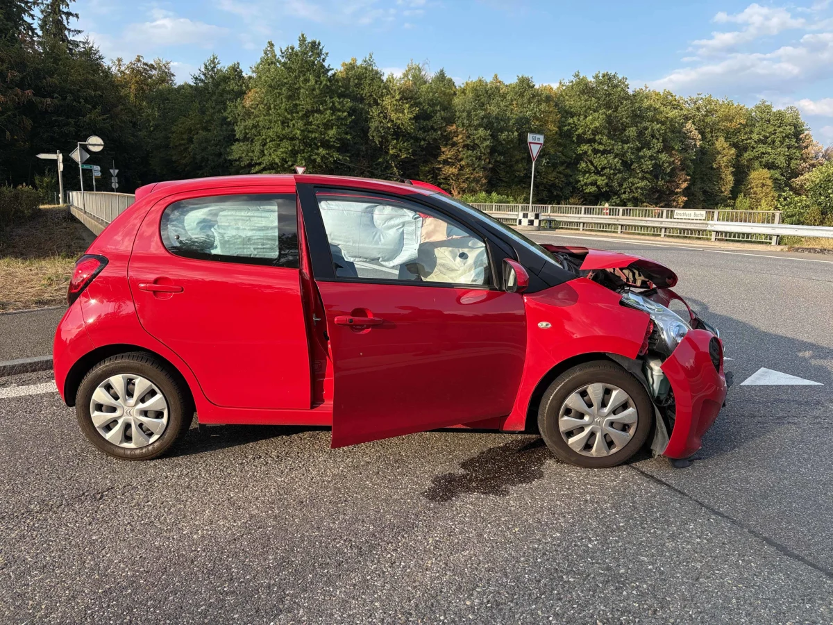 Ein roter Kleinwagen steht mit geöffneter Tür und sichtbaren Airbags am Straßenrand. Die Front ist erheblich beschädigt.