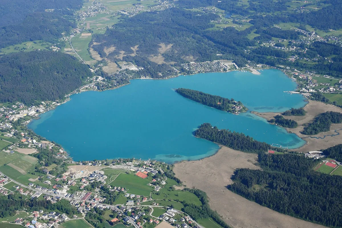 Ein türkisfarbener See ist von oben zu sehen, umgeben von dichten Wäldern und kleinen Siedlungen im Umland. Die Landschaft wirkt friedlich und weitläufig.