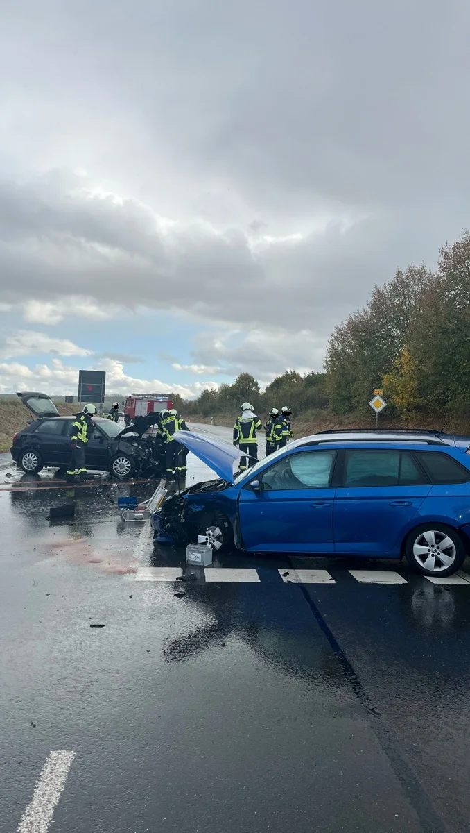 Mehrere Feuerwehrleute sichern die Unfallstelle auf einer nassen Straße, wo zwei Autos sichtbar beschädigt sind. Der Himmel ist bewölkt und es sind Bäume im Hintergrund zu sehen.