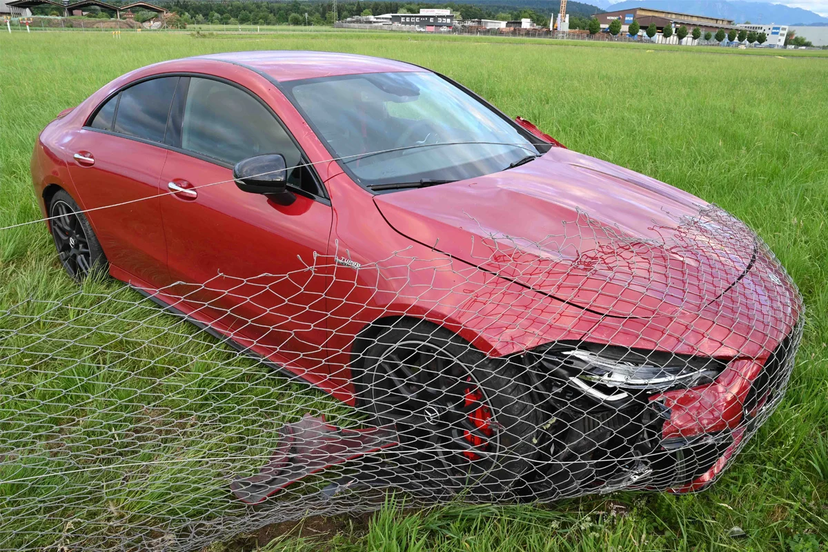 Ein rotes Auto steht in einem Feld und ist durch einen kaputten Drahtzaun beschädigt. Im Hintergrund sind Gebäude sichtbar.