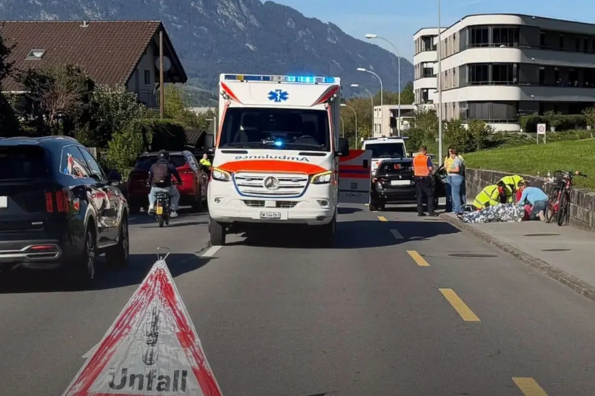 Ein Unfallwagen mit eingeschaltetem Blaulicht steht auf einer Straße. Sanitäter kümmern sich um ein Unfallopfer auf dem Bürgersteig neben Fahrrädern. Polizeibeamte stehen in der Nähe.