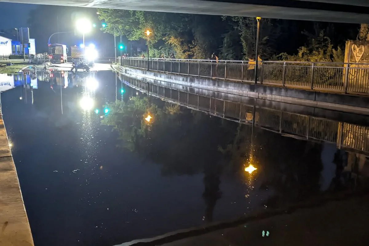 Eine überflutete Straße bei Nacht, beleuchtet von Flutlicht. Einsatzfahrzeuge sind in der Ferne sichtbar.