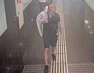 Eine Person trägt eine Decke über der Schulter und geht in einem gefliesten U-Bahn-Tunnel. Die Person trägt dunkle Kleidung und hat kurzes Haar.