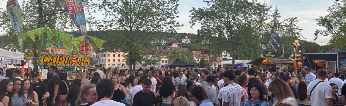 Eine große Menschenmenge versammelt sich bei einem Straßenfest. Verkaufsstände bieten Speisen und Getränke an, und im Hintergrund sind Bäume und Gebäude sichtbar.