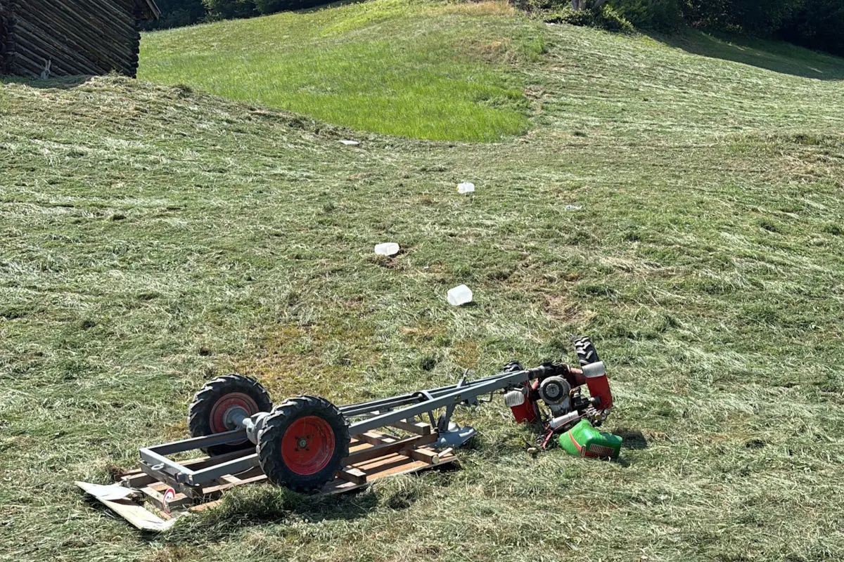 Ein Motormäher liegt umgestürzt auf einer gemähten Wiese. Im Hintergrund ist ein Holzhaufen sichtbar.