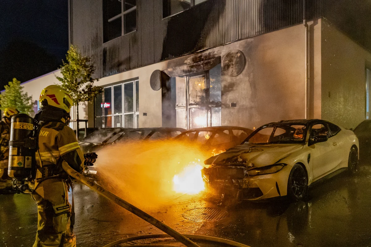 Ein Feuerwehrmann löscht mit einem Wasserschlauch ein brennendes Auto, das vor einem Gebäude geparkt ist. Die Szene findet bei Nacht statt, das Auto ist vom Feuer stark beschädigt.