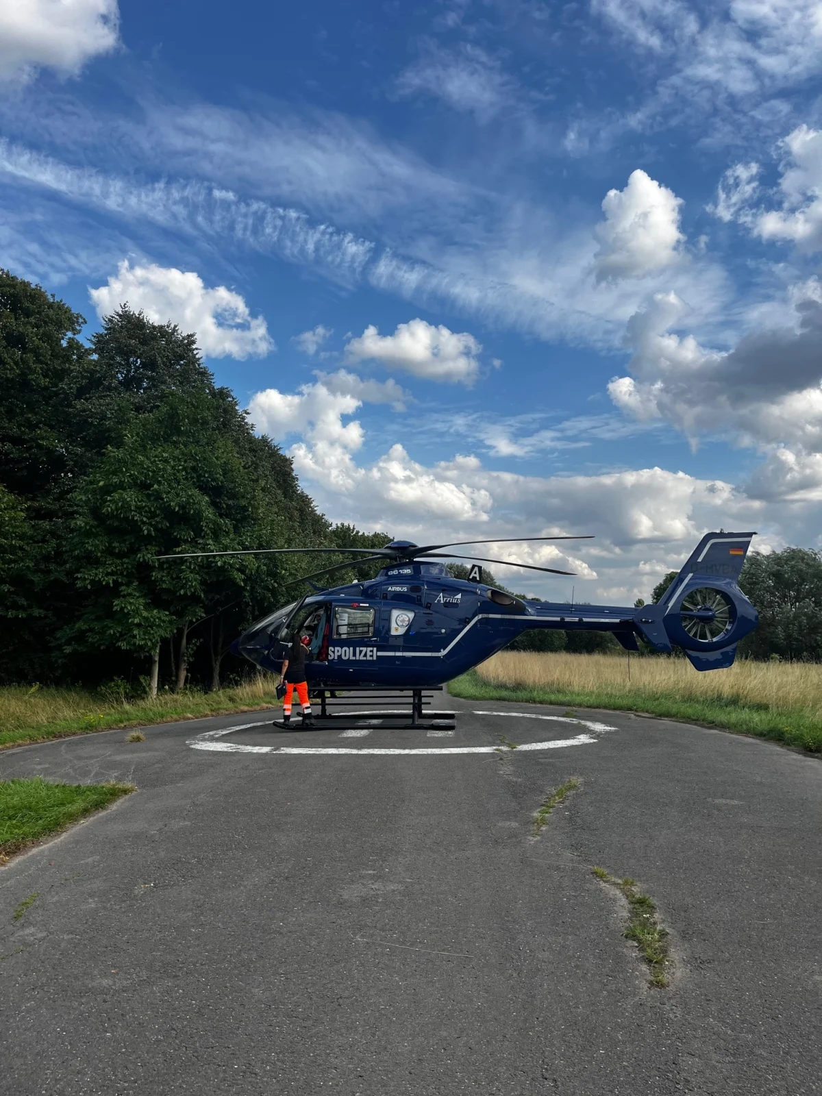 Ein blauer Polizeihubschrauber steht auf einer asphaltierten Landstraße, umgeben von grünen Bäumen und einer Wiese. Ein Mann in orangener Warnkleidung ist neben dem Hubschrauber zu sehen.