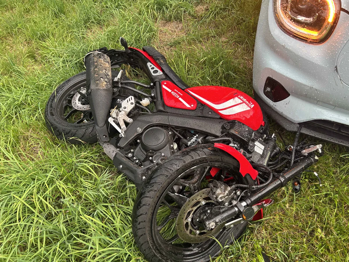 Ein rotes Motorrad liegt umgekippt im Gras neben einem grauen Auto. Die Fahrzeuge sind nass vom Regen.