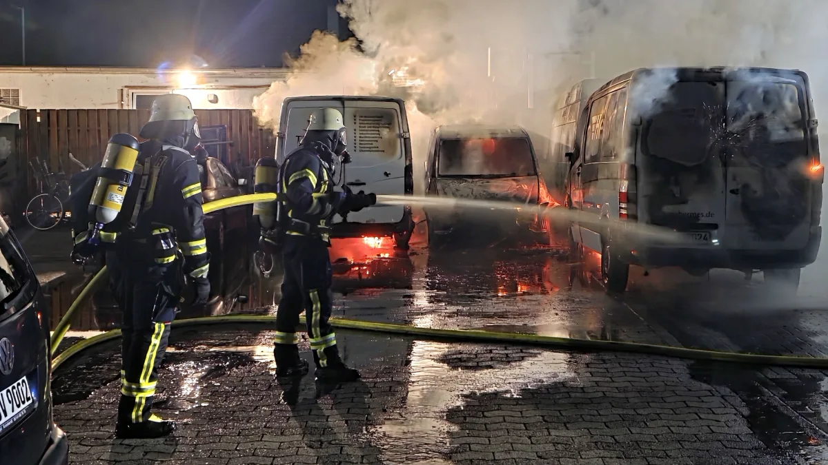Zwei Feuerwehrleute in voller Ausrüstung löschen mit einem Schlauch brennende Transporter auf einem Parkplatz. Dichter Rauch umhüllt die Szene.