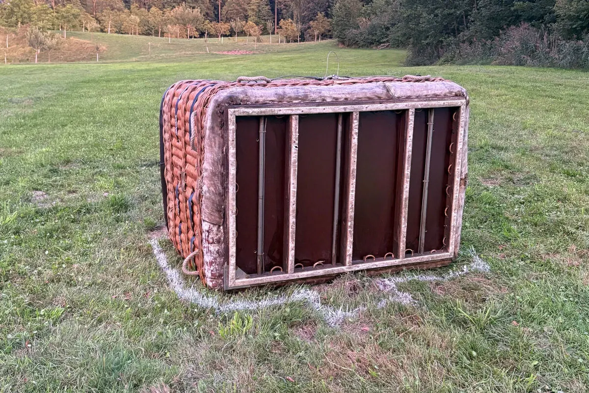 Ein umgekippter Heißluftballonkorb liegt auf einer Wiese; der Korb ist aus Weidengeflecht und innen mit Metall verstärkt.