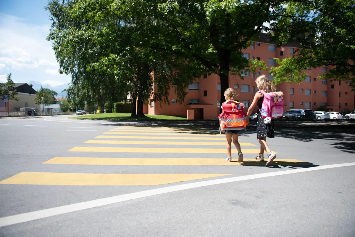 Der vordere Bereich zeigt zwei Kinder mit Schulranzen, die bei Tageslicht einen Zebrastreifen überqueren. Im Hintergrund sind Wohngebäude und Bäume zu sehen.
