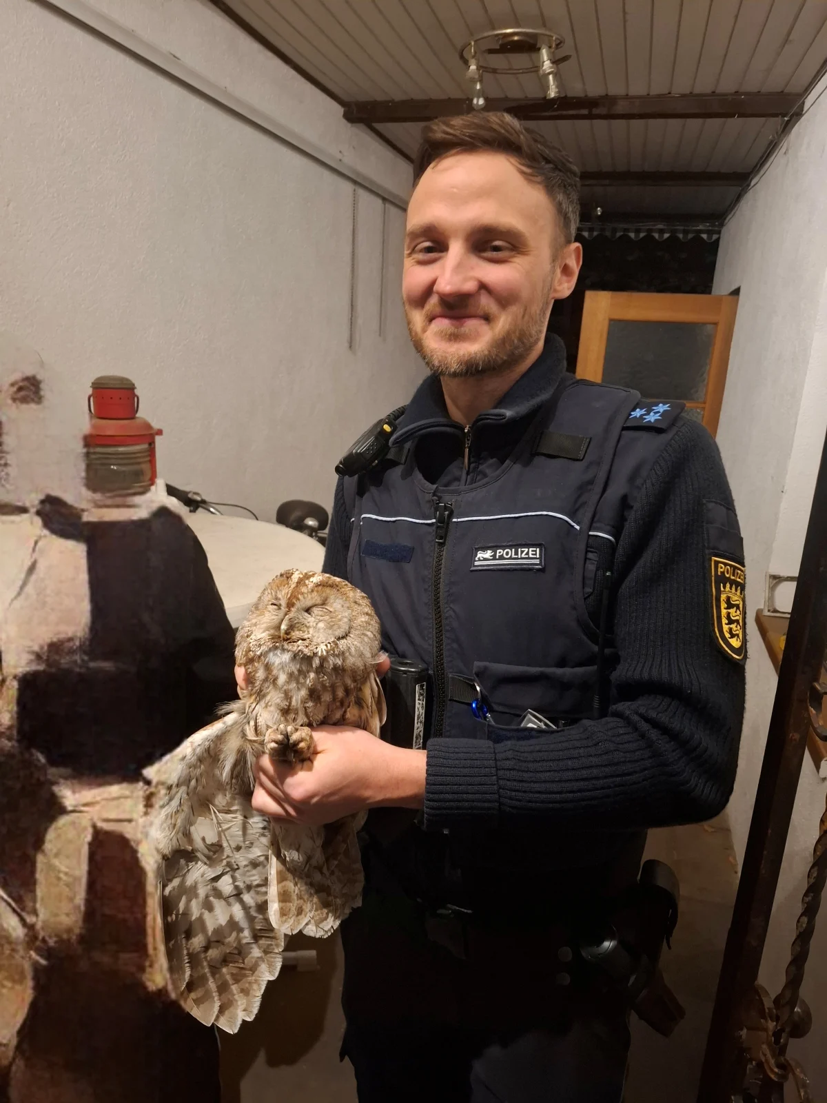 Ein lächelnder Polizist in Uniform hält eine Eule in einem Wohnraum. Im Hintergrund sind verschiedene Gegenstände zu sehen.