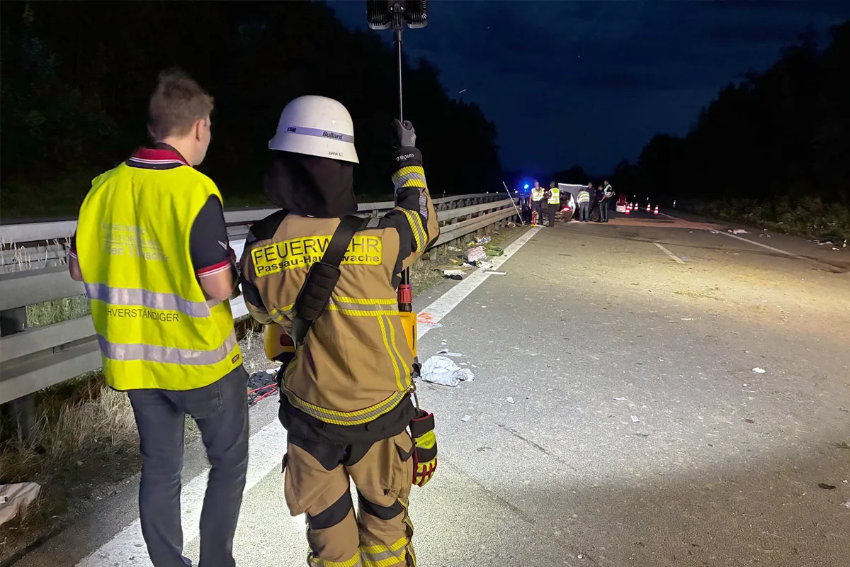 Ein Feuerwehrmann in Schutzkleidung hält eine Lampe an einem nächtlichen Unfallort auf einer Autobahn. Ein weiterer Mann in einer Warnweste steht daneben, während im Hintergrund weitere Einsatzkräfte bei einem Fahrzeug arbeiten.