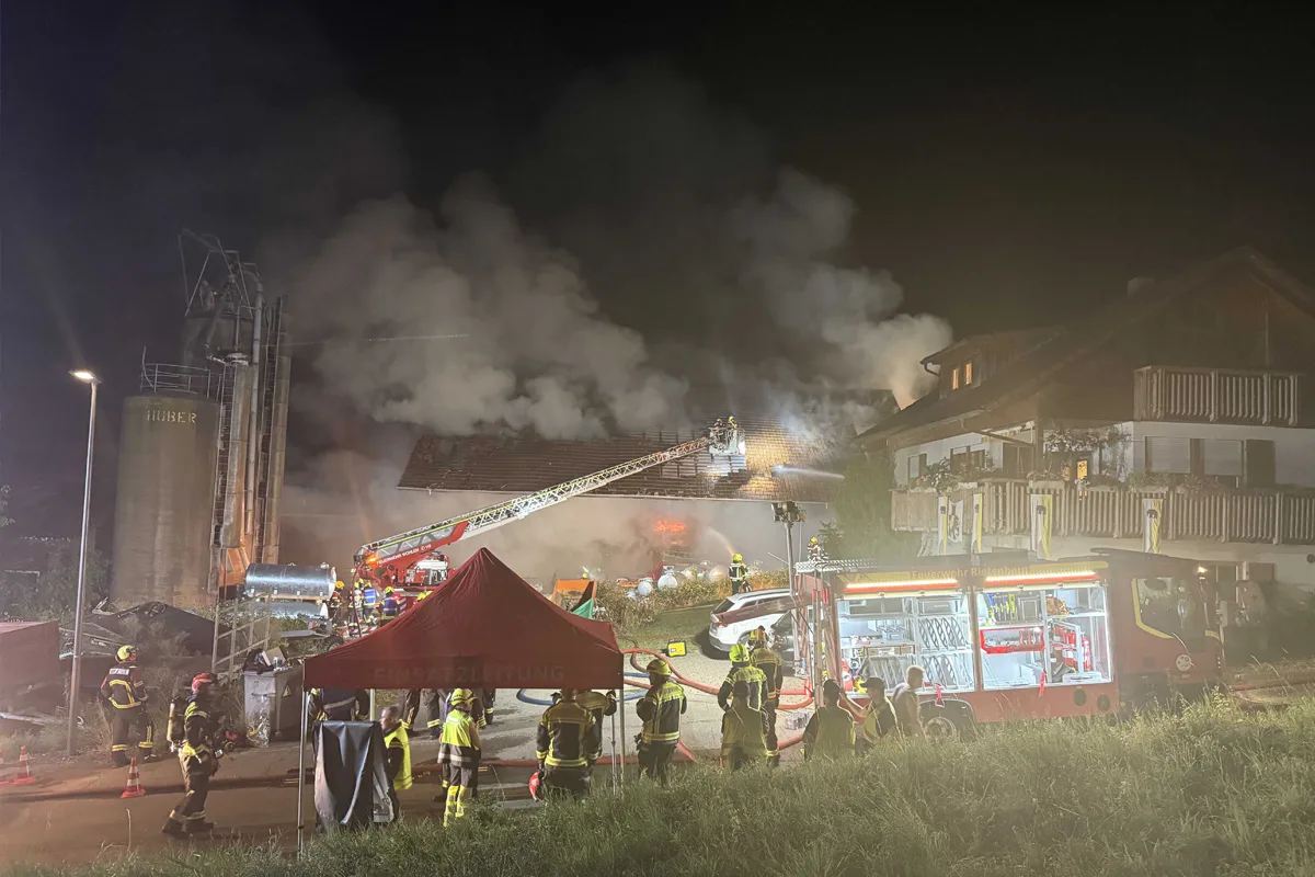 Feuerwehrleute sind bei Nacht im Einsatz, um einen Brand auf einem Dach zu löschen. Mehrere Feuerwehrfahrzeuge und ein Einsatzzelt sind vor Ort.