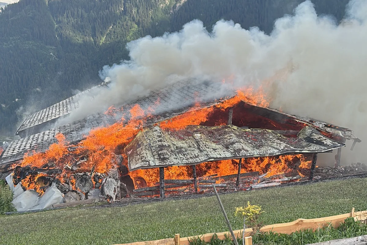 Große Flammen und dichter Rauch steigen von dem brennenden Gebäude auf, das in einer bergigen Umgebung liegt.