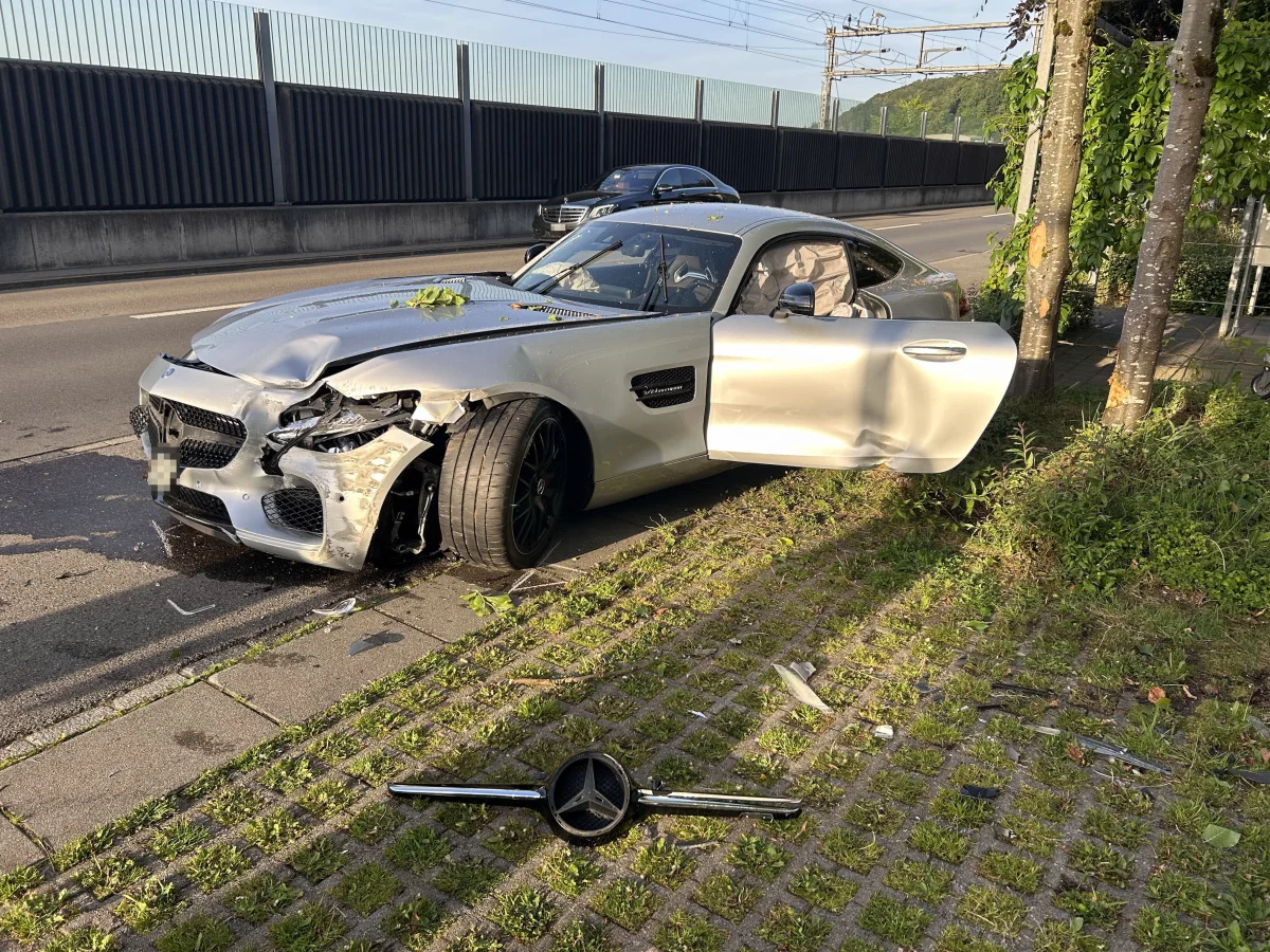 Ein silbernes Auto mit erkennbaren Frontschäden steht am Straßenrand. Die Fahrertür ist geöffnet, und einzelne Autoteile liegen auf dem Boden.