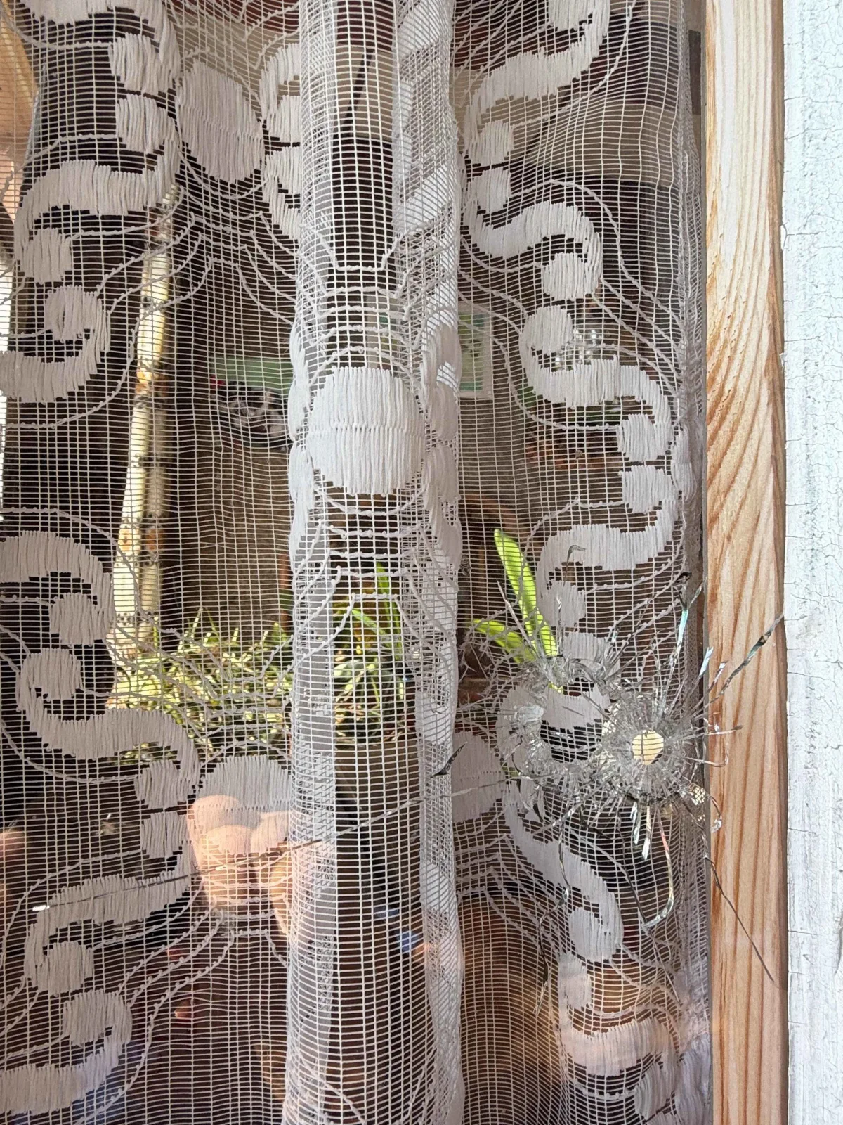 Ein Fenster ist beschädigt durch ein Einschussloch, das hinter einer weißen Spitzengardine sichtbar ist.