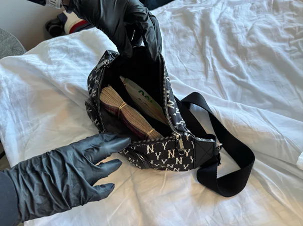 Eine Person mit schwarzen Handschuhen öffnet eine schwarz-weiße Tasche, in der mehrere Geldbündel sichtbar sind. Die Tasche liegt auf einem weißen Bettlaken.