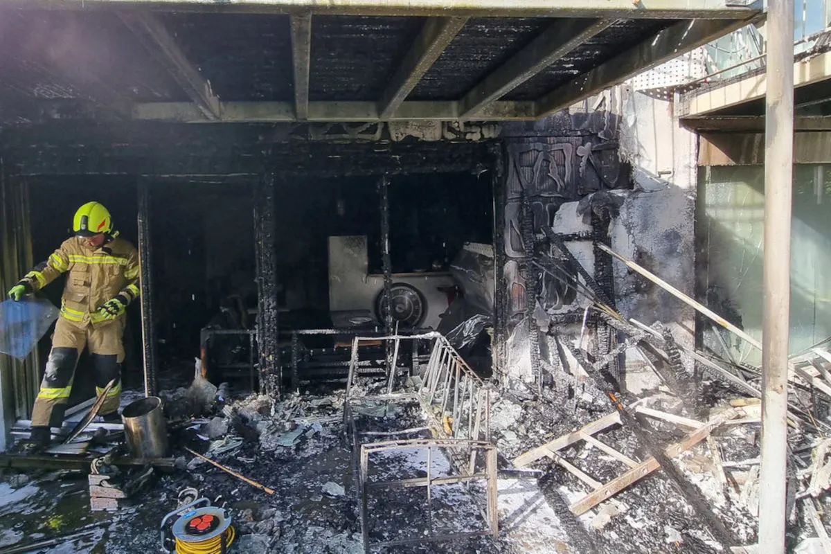 Ein Feuerwehrmann in voller Einsatzkleidung betrachtet die verkohlten Überreste eines Gebäudes. Um ihn herum liegen Trümmer und verbranntes Material.