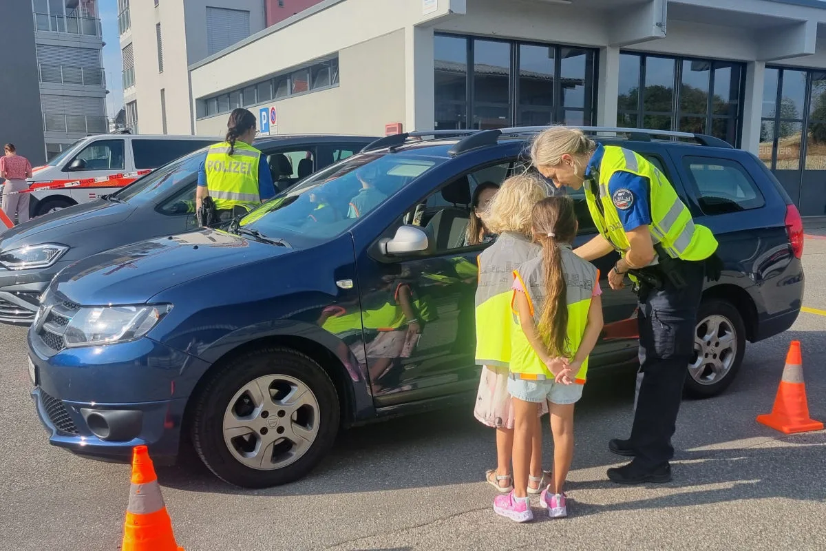 Eine Polizistin in gelber Weste spricht mit zwei Kindern in Sicherheitswesten neben einem blauen Auto. Im Hintergrund sind weitere Fahrzeuge und ein modernes Gebäude zu sehen.