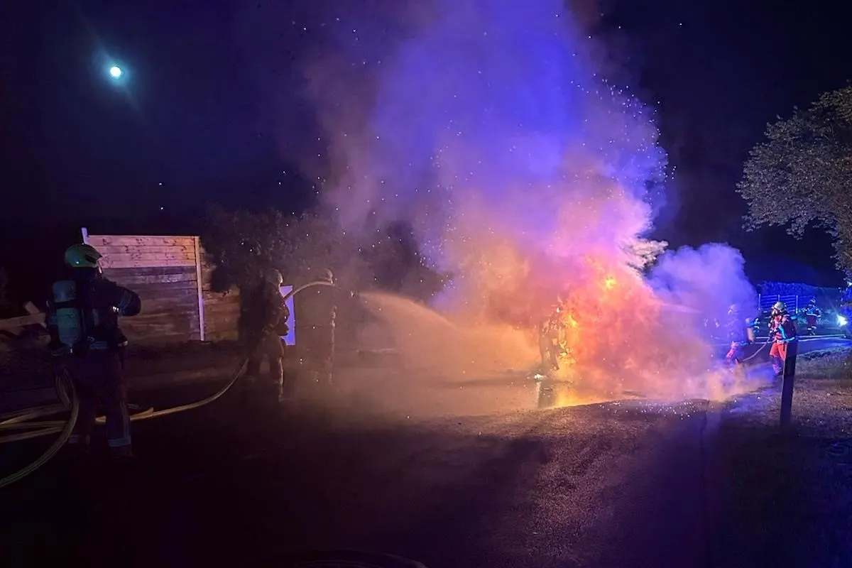 Mehrere Feuerwehrleute sind zu sehen, wie sie bei Nacht mit Wasserschläuchen ein stark brennendes Fahrzeug löschen. Das Bild ist von Rauch und leuchtenden Flammen geprägt.