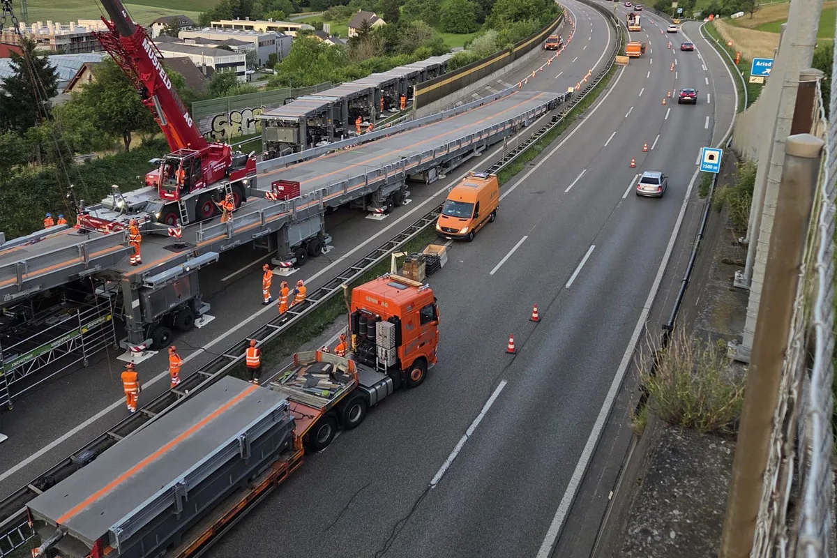 Mehrere Arbeiter in leuchtenden Warnwesten sind auf einer temporären Brückenkonstruktion aktiv; ein roter Kran ist im Einsatz, während Fahrzeuge die Autobahn passieren.