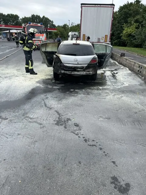 Ein stark beschädigtes, ausgebranntes Auto steht mit geöffneten Türen auf einem Rastplatz. Ein Feuerwehrmann steht neben dem Fahrzeug, auf dem Boden sind Spuren von Löschmitteln sichtbar.