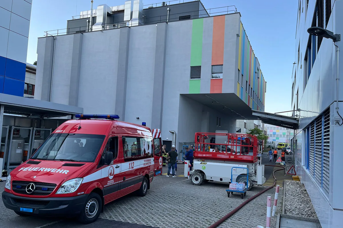 Ein roter Feuerwehrwagen parkt vor einem bunten, modernen Gebäudekomplex. Feuerwehrpersonal ist ebenfalls sichtbar.